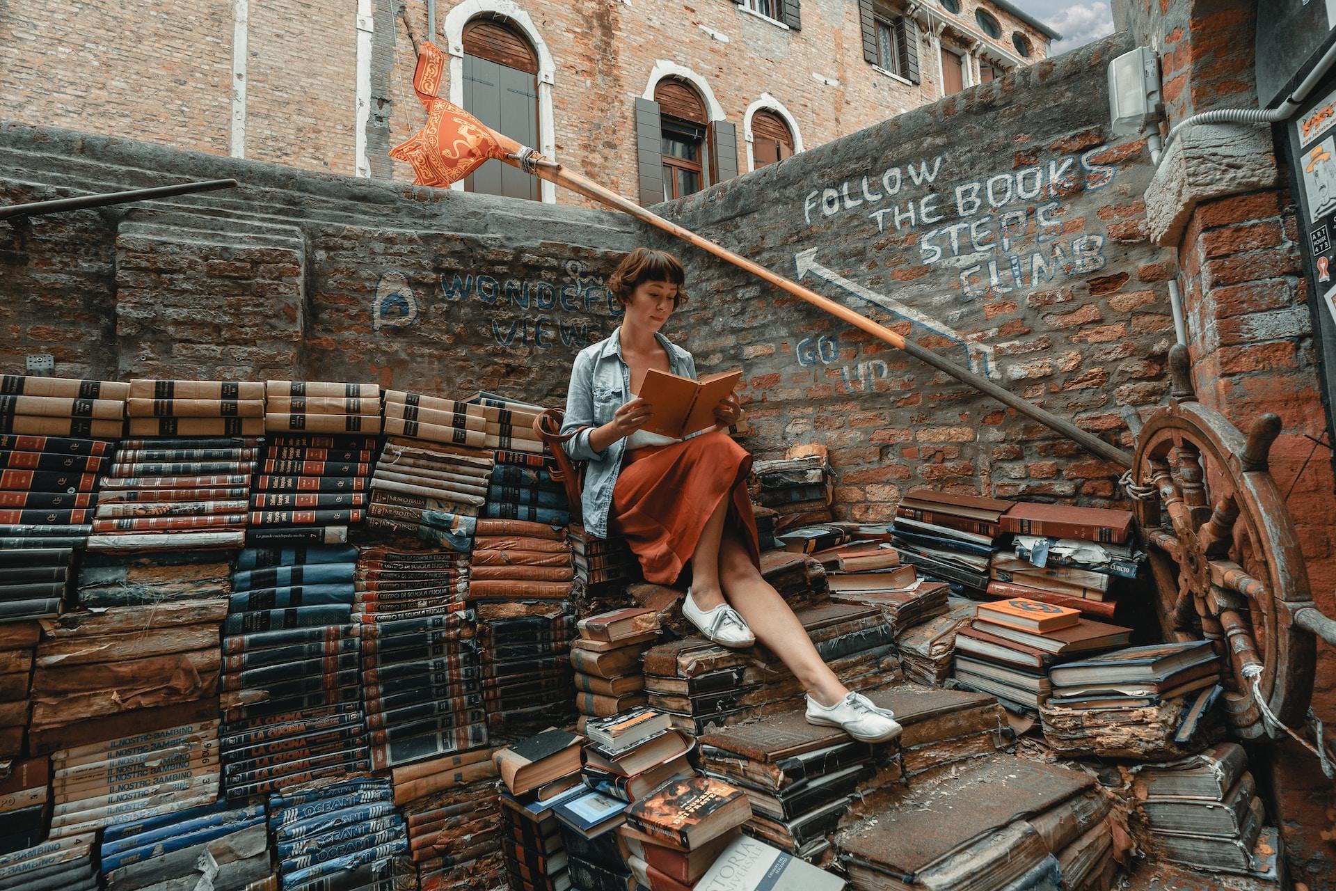 A girl sits on an extremely large pile of stacked books in an alleyway. She is reading one of the books.