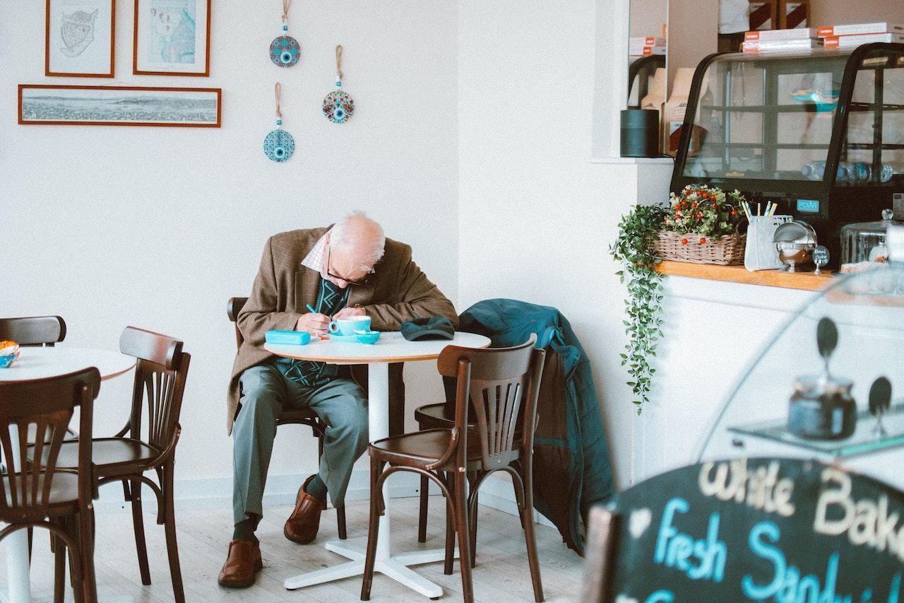An older man sits at a table in a bright cafe writing in a notebook.