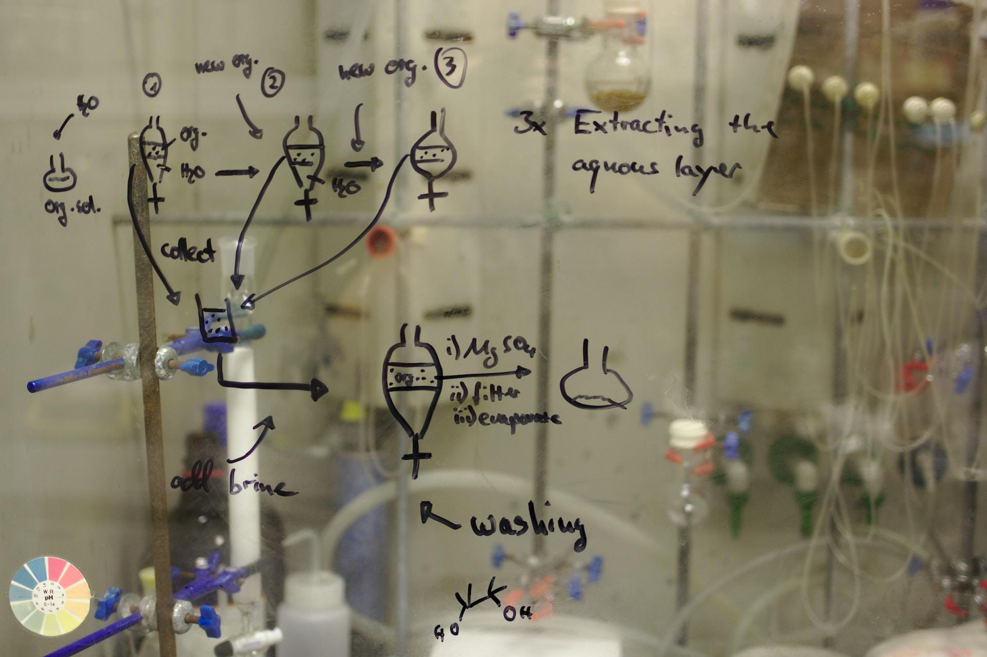 A transparent dry-erase board displays various chemical processes. Through the board, you can recognise the trappings of a chemistry lab.