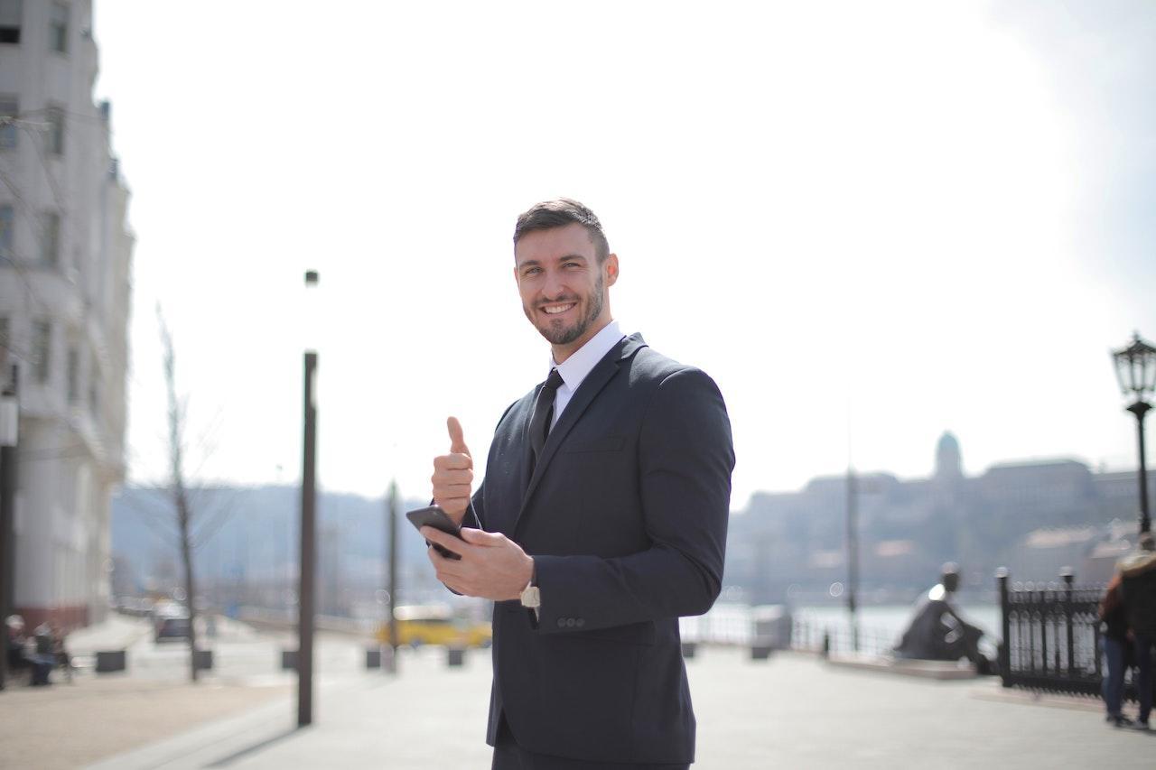 A man in a business suit gives a thumbs up in front of a European city.
