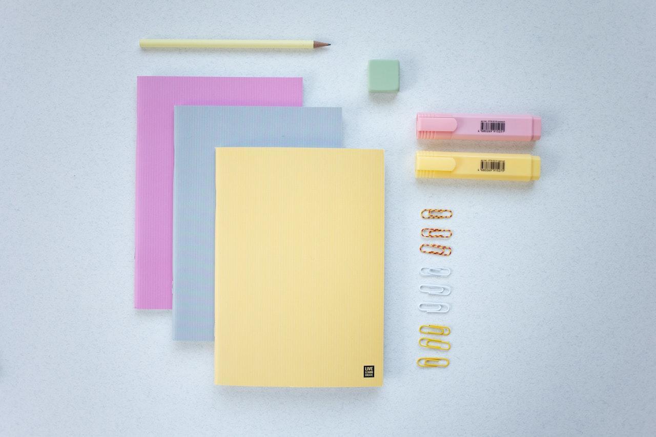Notebooks, a pencil, an eraser, highlighter markers, and paperclips arranged neatly on a white desk.
