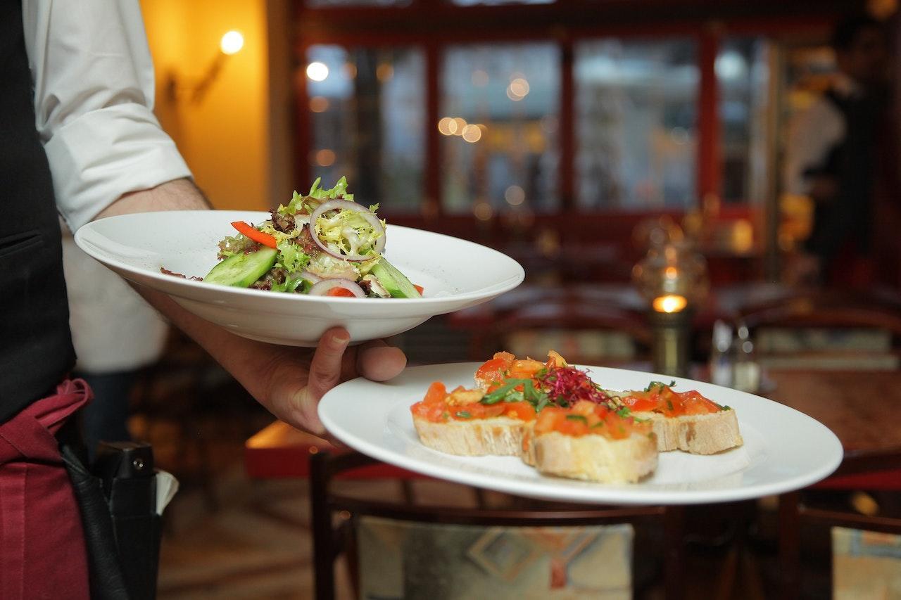 A waiter holds two plates of fine dining styled food: a salad and a scallop dish.