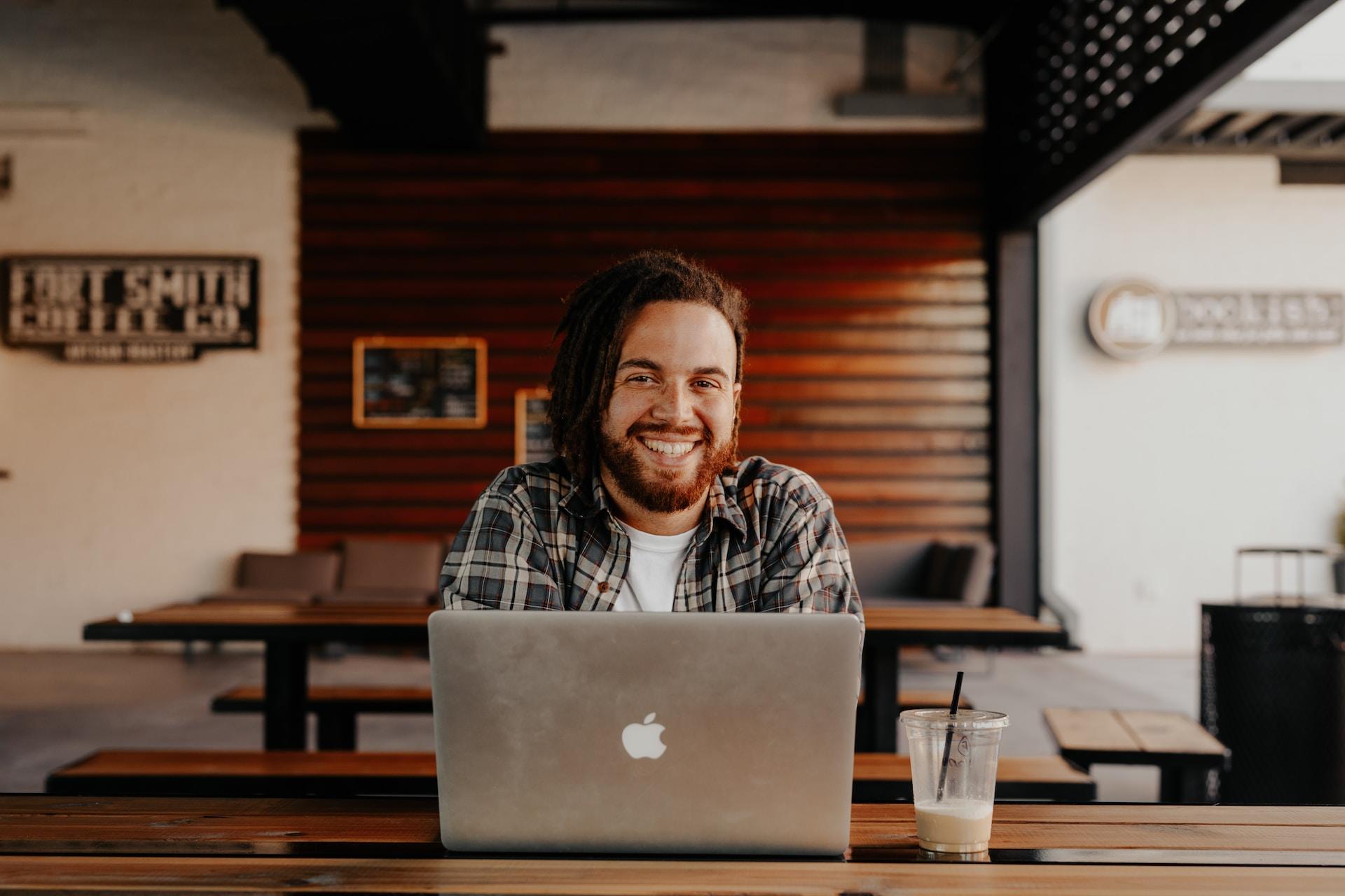 A smiling bearded man with longish red hair sits in a cafe with a half-drunk plastic cup of beverage beside his open laptop computer.
