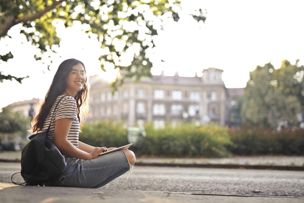 A girl sits cross-legged on a stone path outside with an historic stone building in the background.