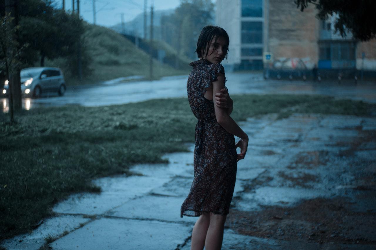 A girl stands outside on a dark, rainy day with her dress plastered to her body, looking backwards over her shoulder at the camera.