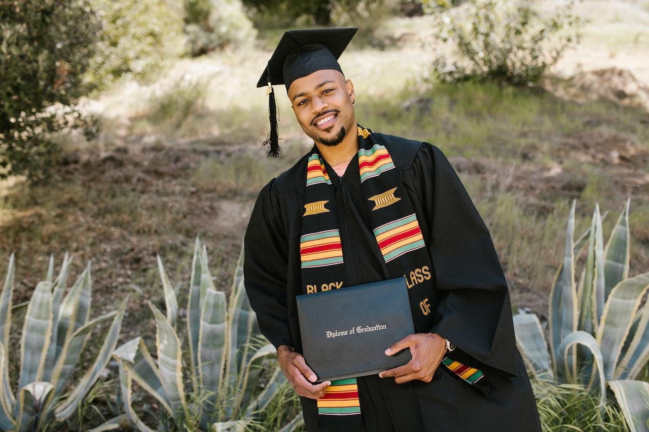 A young man stands outside in front of a desert scrub background dressed in a black graduation cap and robe. He has a brightly-patterned stole and is holding his diploma.