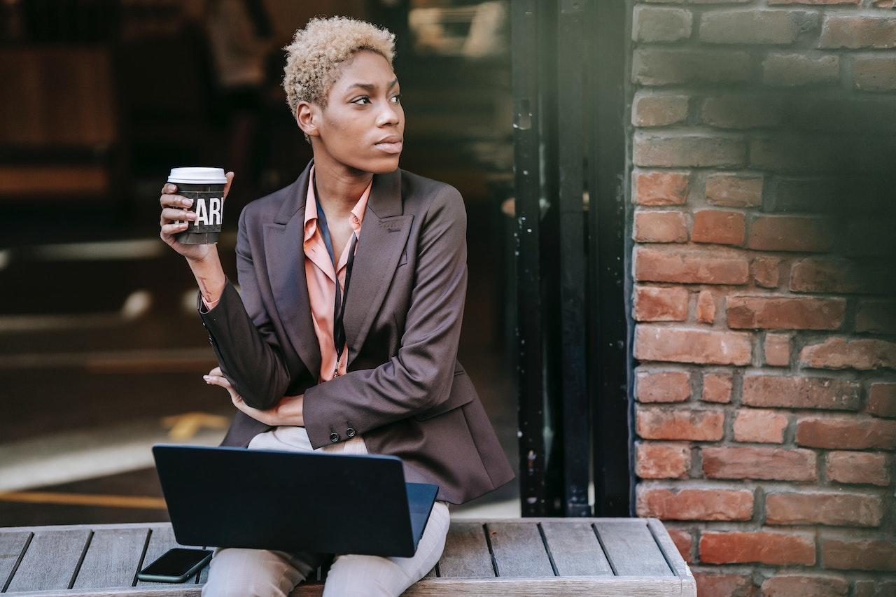A person sits on a wooden bench outside of a shop holding a paper cup of coffee with a laptop on their lap.
