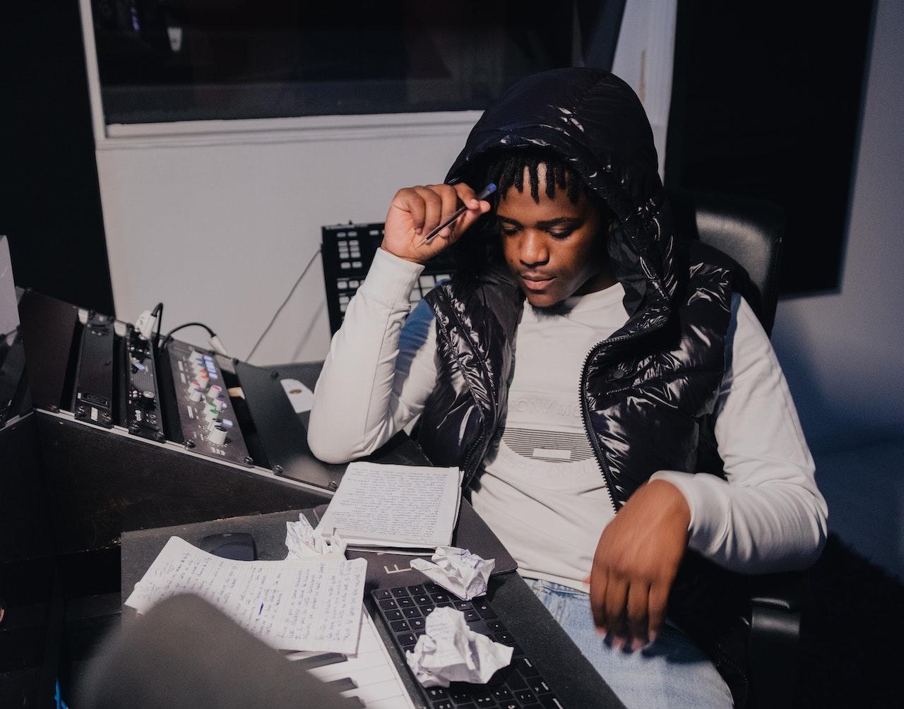 A man sitting in a sound booth writes lyrics in a notebook. There are crumpled papers on the desk as well.