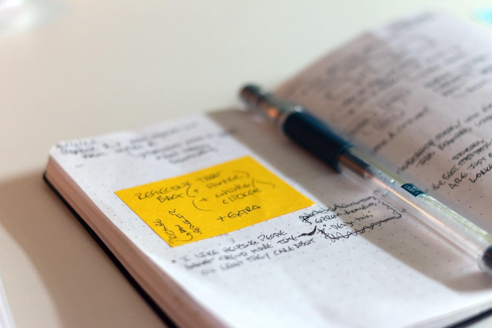 A close up of a page in a notebook with handwriting and a yellow sticky note with a pen resting on the paper.