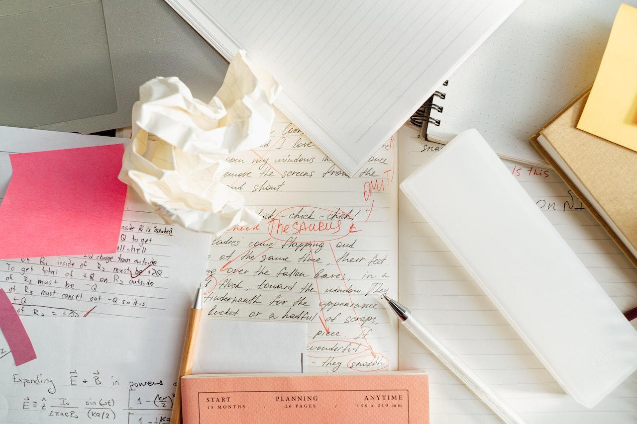 A mishmash of papers are on a desk, and the center paper has a lot of writing on it as well as some red pen remarks about improving the writing.