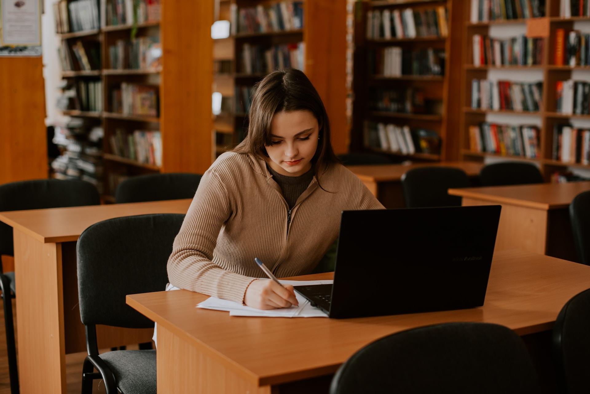 A person in a light brown sweater sits at a table in a library with an open laptop in front of them. But they write notes by hand.