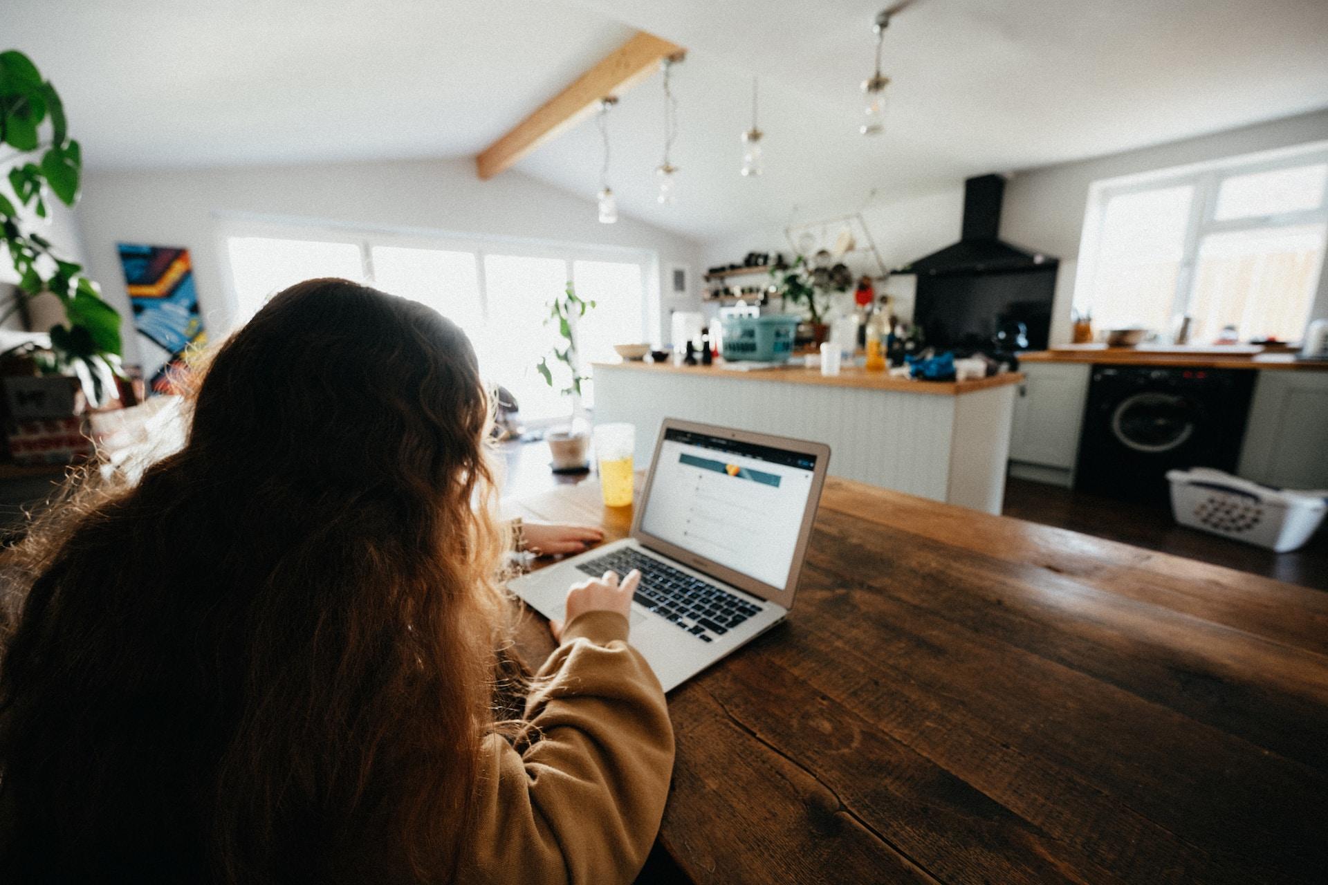 A person with long hair wearing a brown sweater sits at a kitchen table, facing the cooking area. There is a laptop computer open in front of them and they are working the touchpad while looking at the screen.