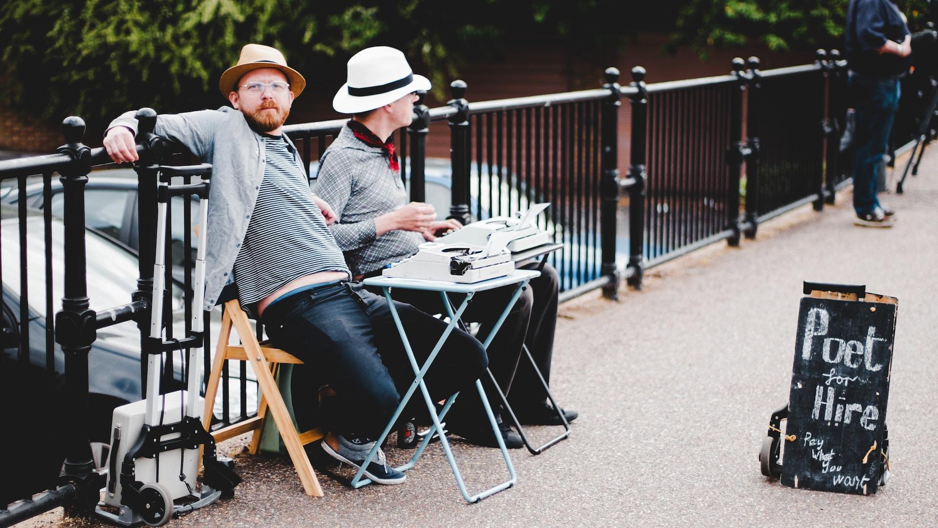 Two men sit at a small table outside on the sidewalk with a typewriter. A sandwich board sign in front of them reads "Poet for hire. Pay what you want."