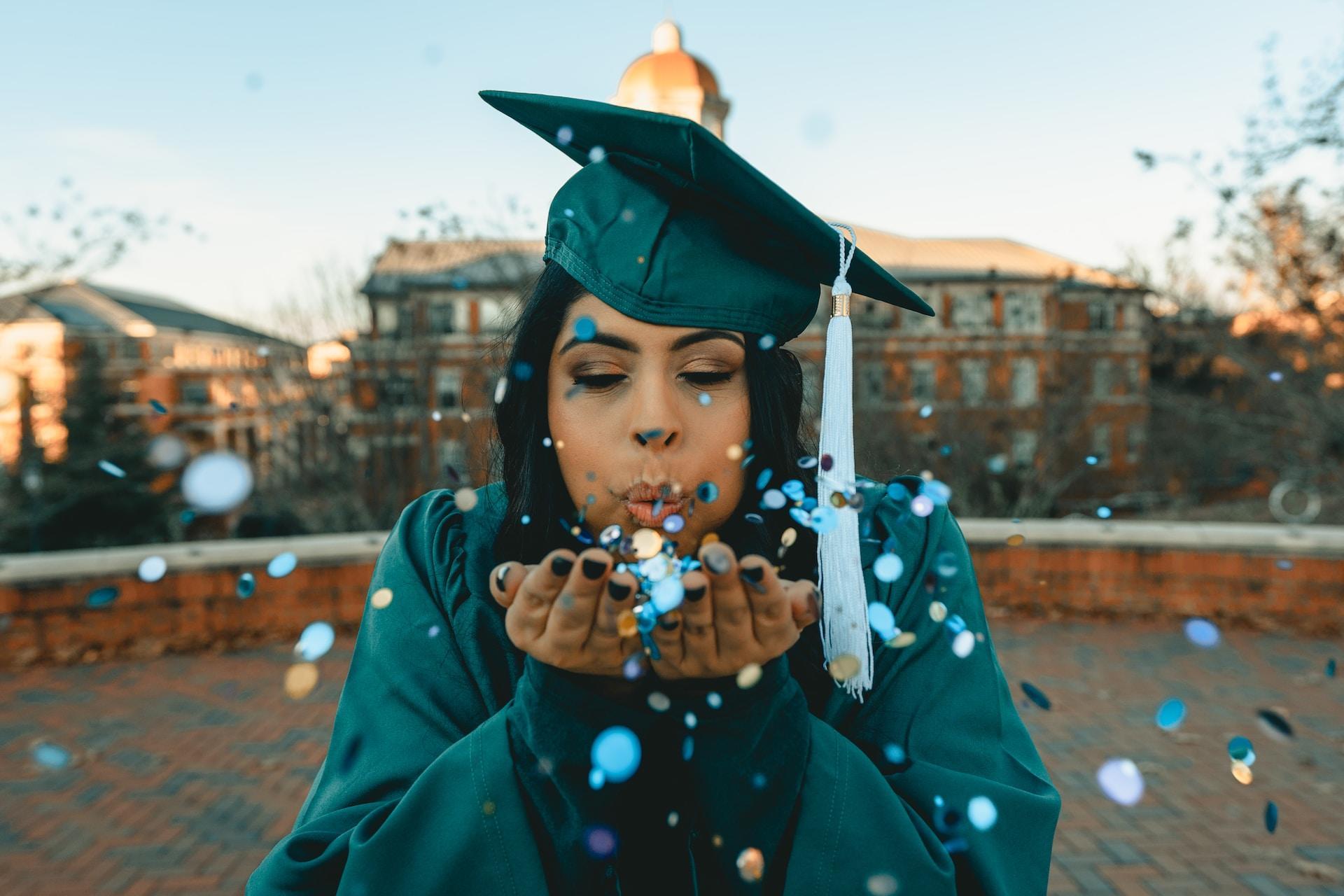 A graduate in a cap and gown blows blue glitter out of her cupped hands towards the camera.