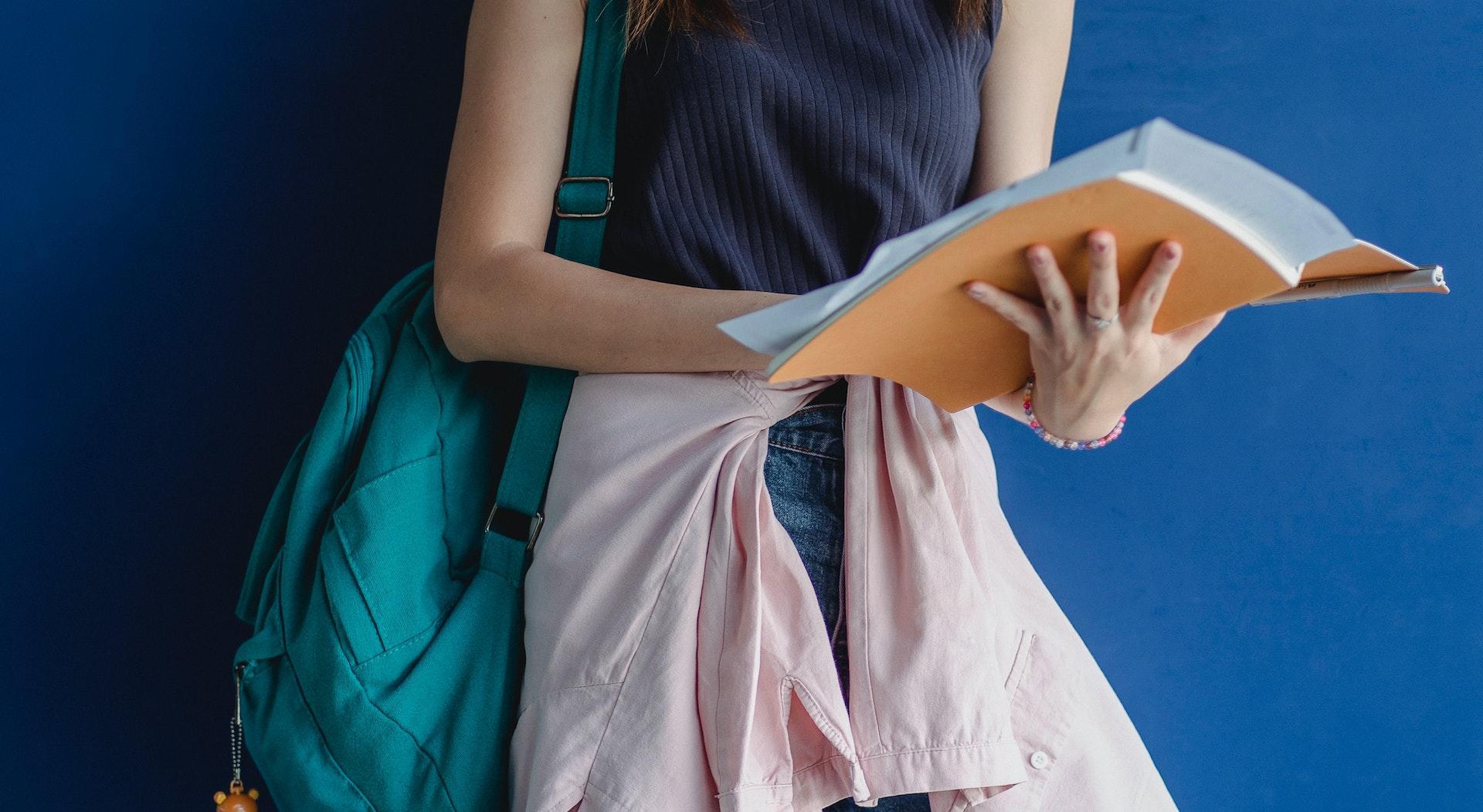 A student stands against a bright blue wall holding an open textbook with paper resting on the page.