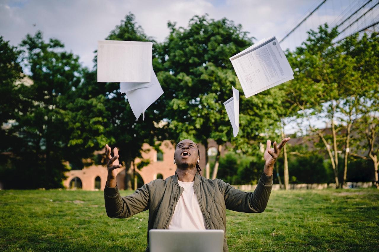 A student sitting outside on grass throws his notebooks into the air