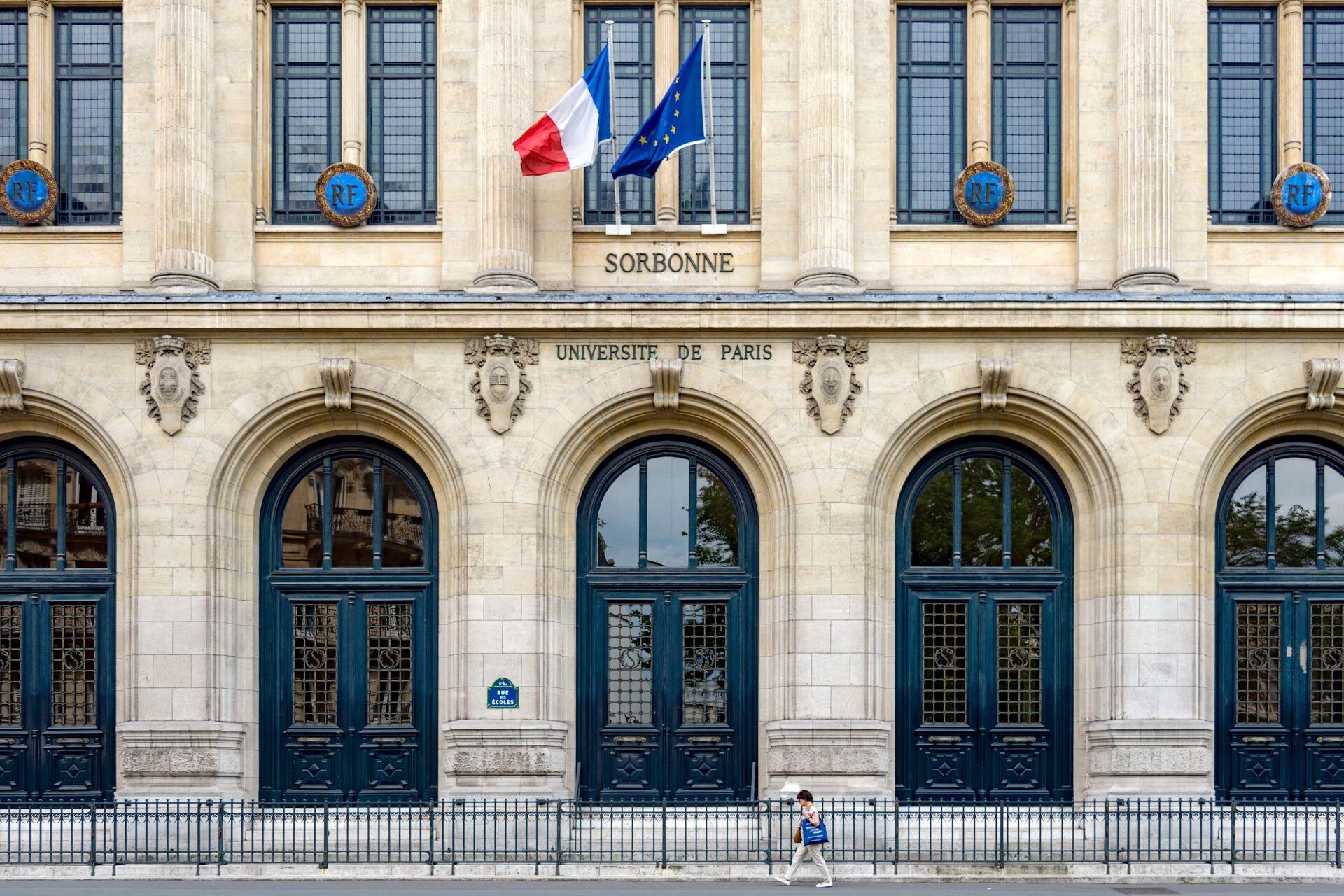 Stone façade of the Sorbonne with arched doorways and French and EU flags
