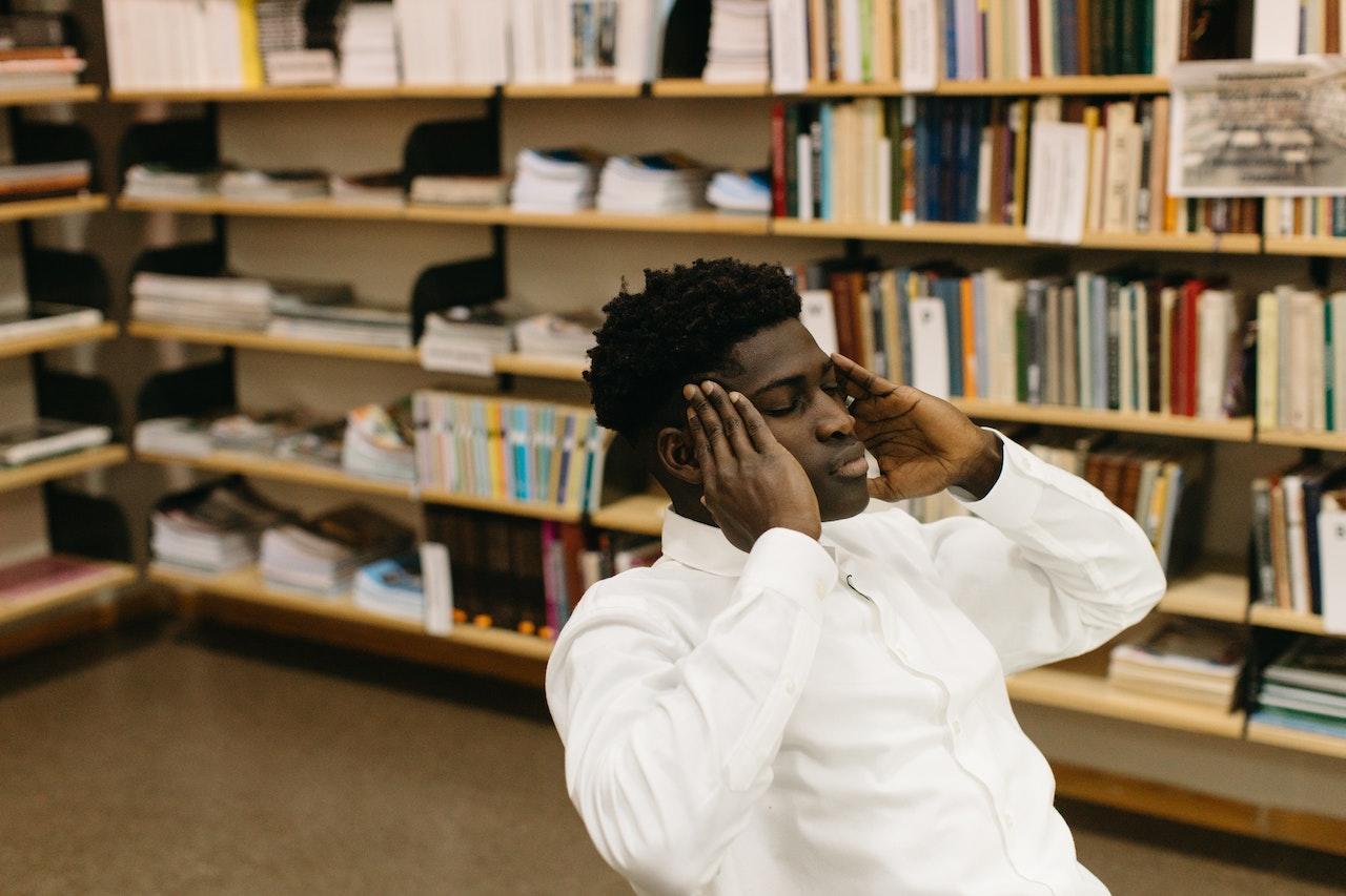 A student sits in the library with his hands on his temples and eyes closed, thinking