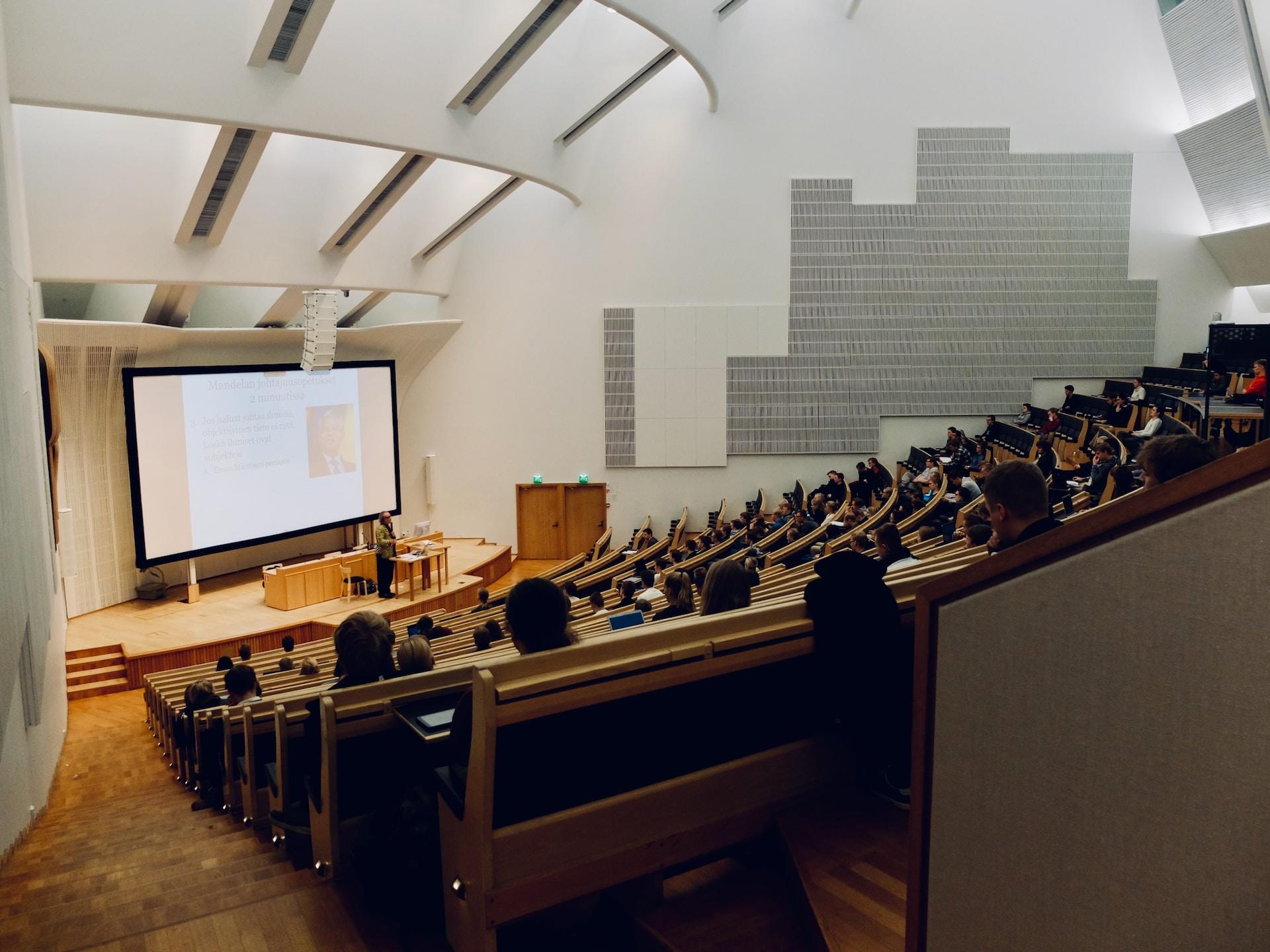 A university lecture hall outfitted with light-coloured wood fixtures against a white decor, seen from the uppermost tier, looking down at the presentation screen.