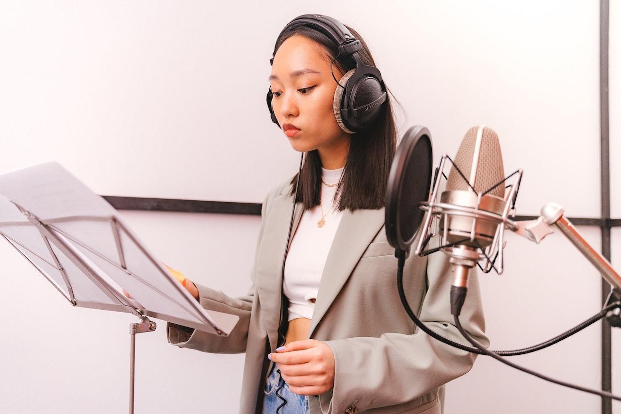 A woman wearing headphones in a recording booth looks at a script on a music stand. A microphone with windscreen is suspended nearby.