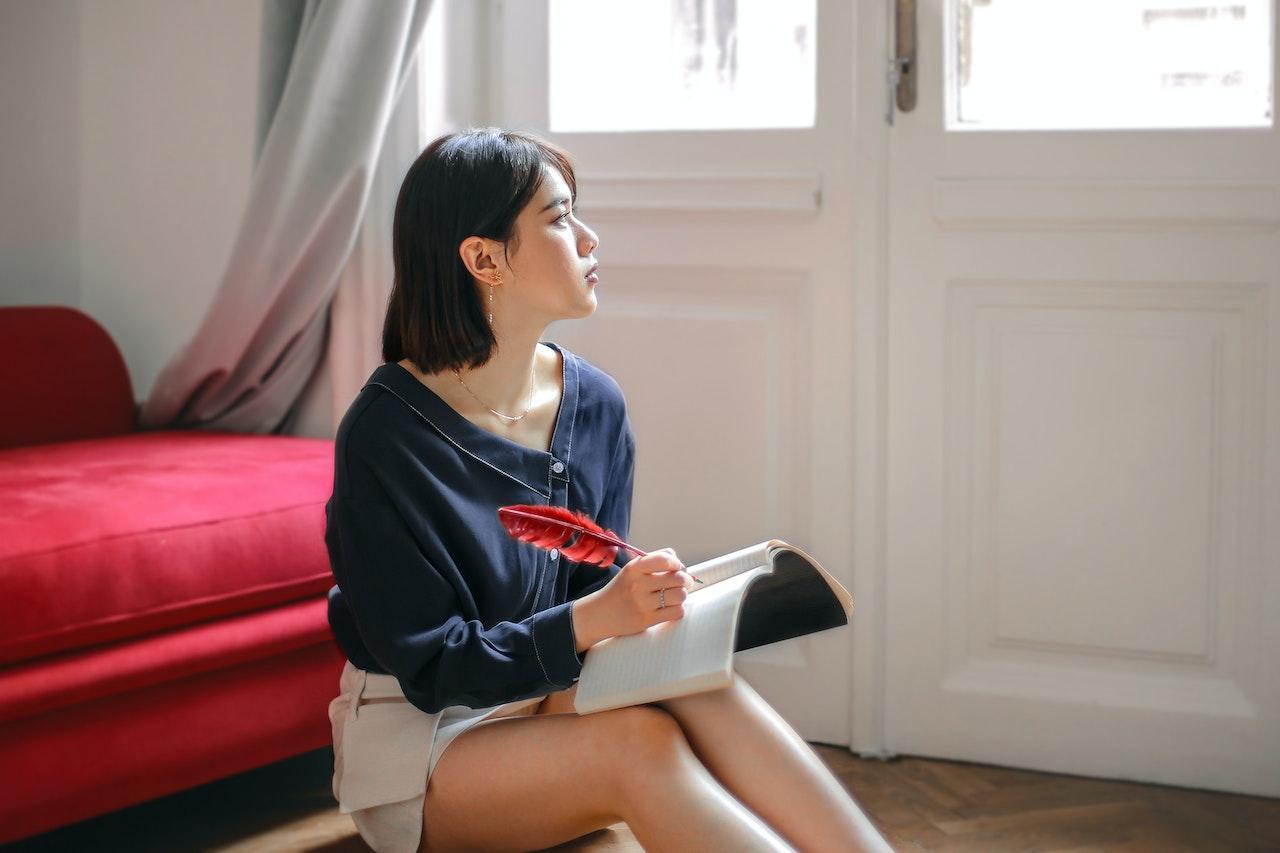 A girl sitting on a floor pouf in a simple room holds a notebook and a feather quill pen while looking out the window wistfully.