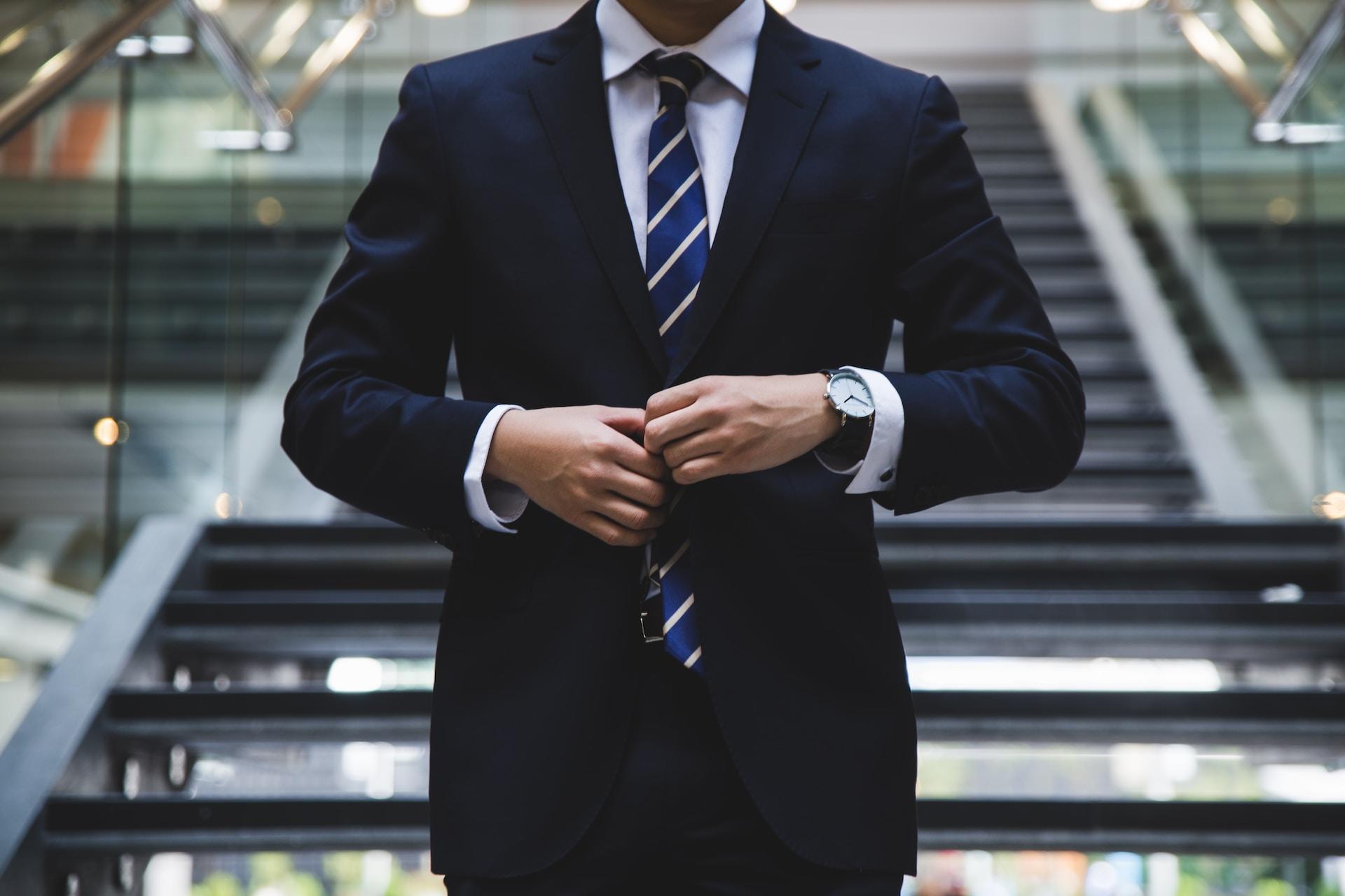 A person in a dark blue business suit over a light blue shirt and striped tie buttons their jacket while standing in front of a wide staircase with shallow stairs.