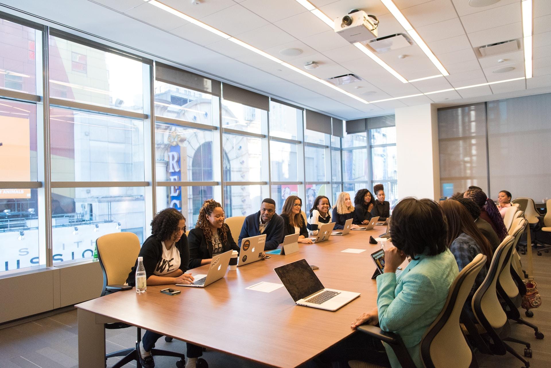 A large group of people wearing business attire sit around a light-coloured wooden table, some with open laptops in front of them, in a room with a bank of windows letting in natural light. 