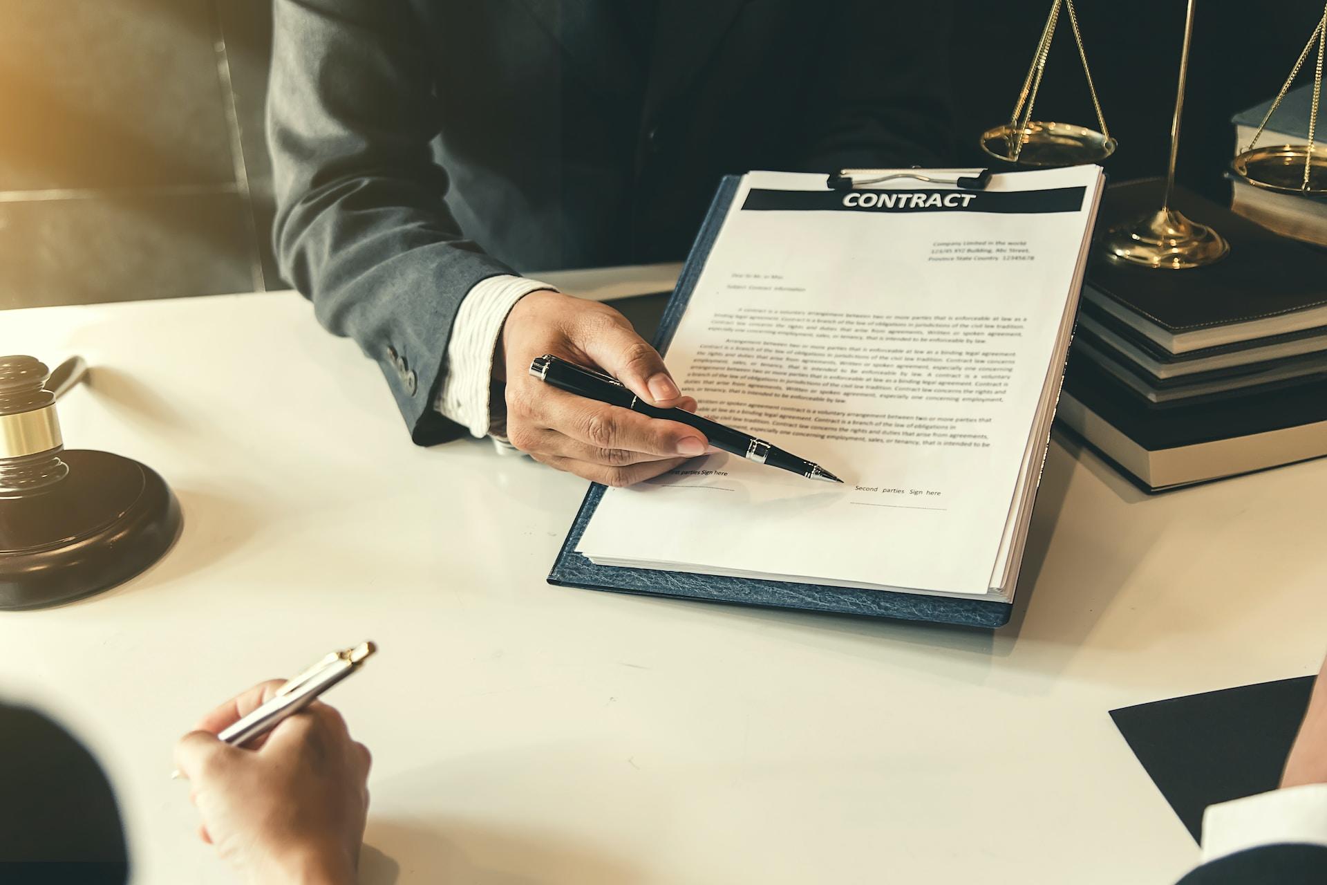 A person wearing a business suit tilts a clipboard bearing a contract towards a person sitting on the other side of the table with a pen in their hand, ready to sign where the first person is pointing. 