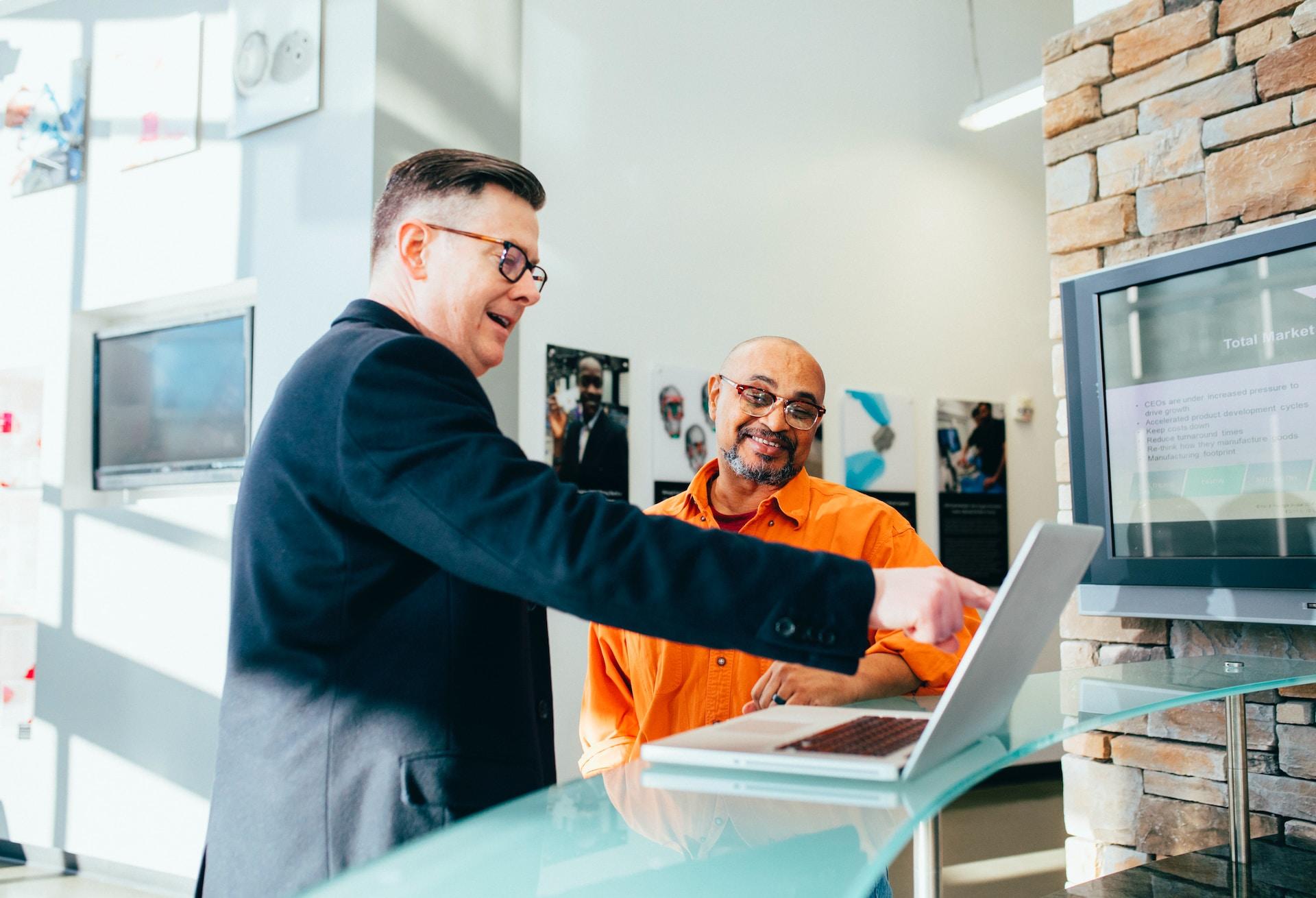 A person wearing a dark coloured suit stands and points to an open laptop's monitor while a person wearing an orange shirt and glasses looks on and smiles. 
