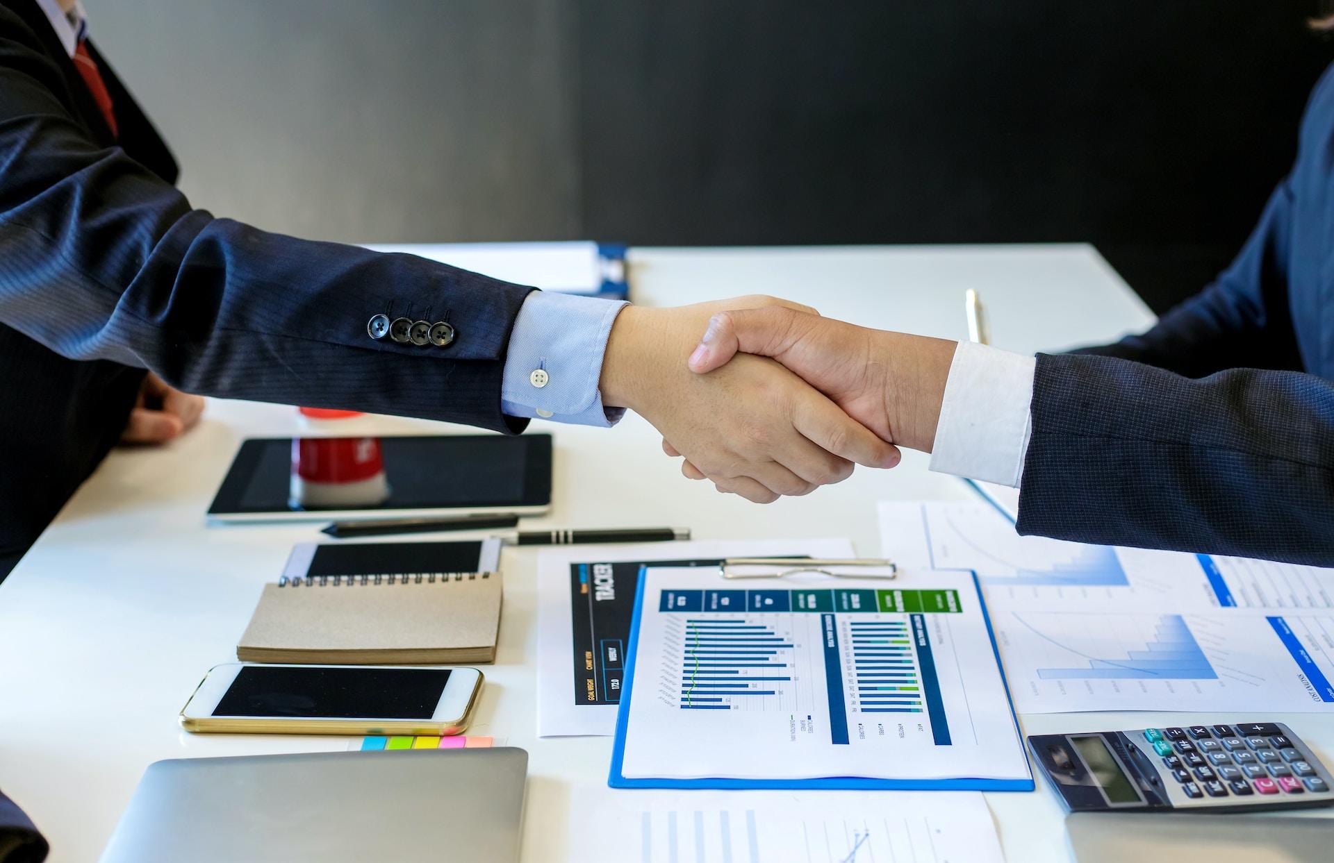 Two people wearing dark business suits reach across a table laden with paperwork and charts to shake hands. 
