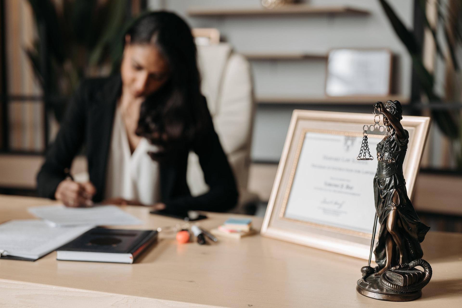 A lawyer makes notes on her desk while a statuette of Lady Justice is on the front of the desk in the foreground.