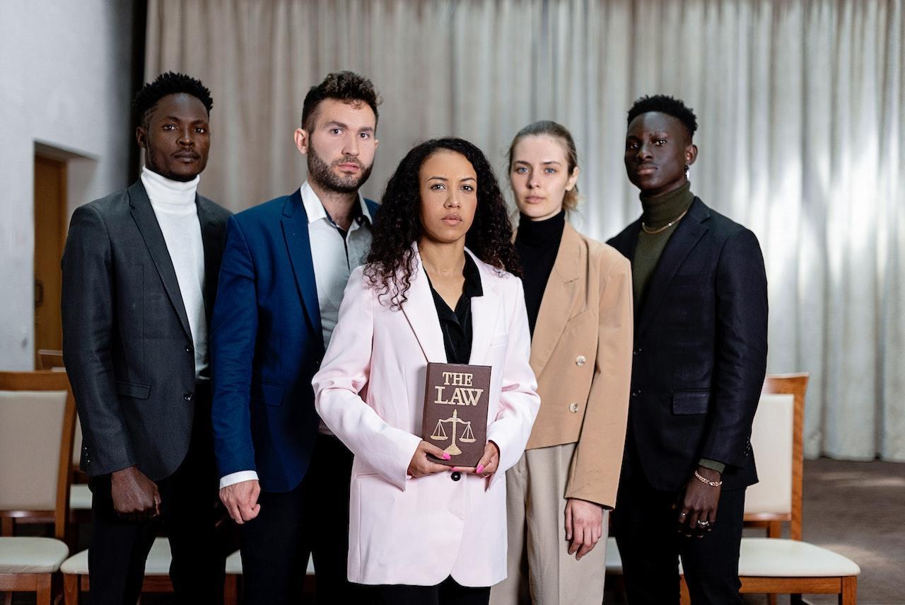 A group of lawyers stand together in an office. The one in the front holds a book titled "The Law."