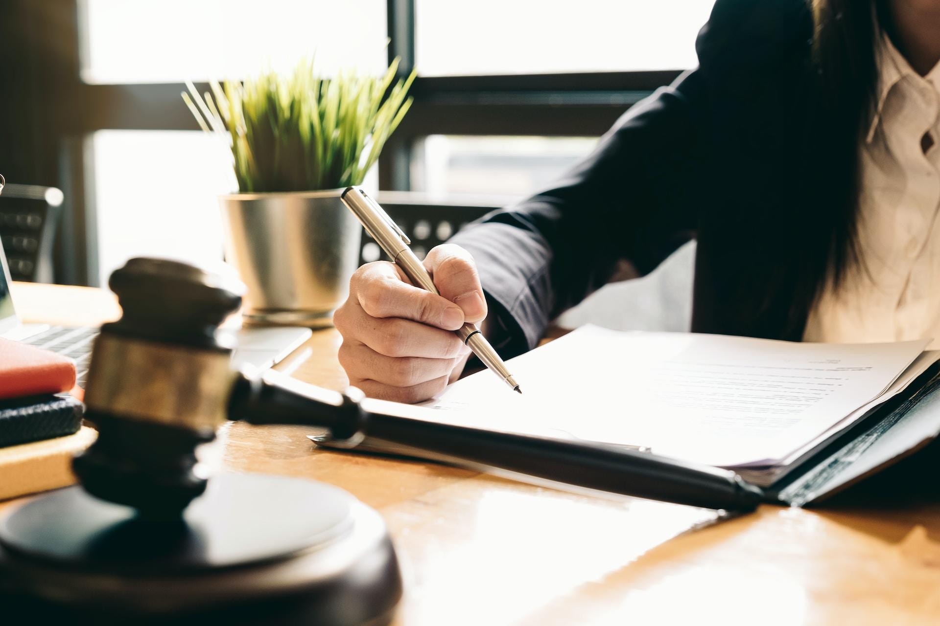 A person wearing business attire holds a pen in their right hand, preparing to sign a document. In front of them, on their desk, rests a gavel and next to them, in the background, is a small green plant.