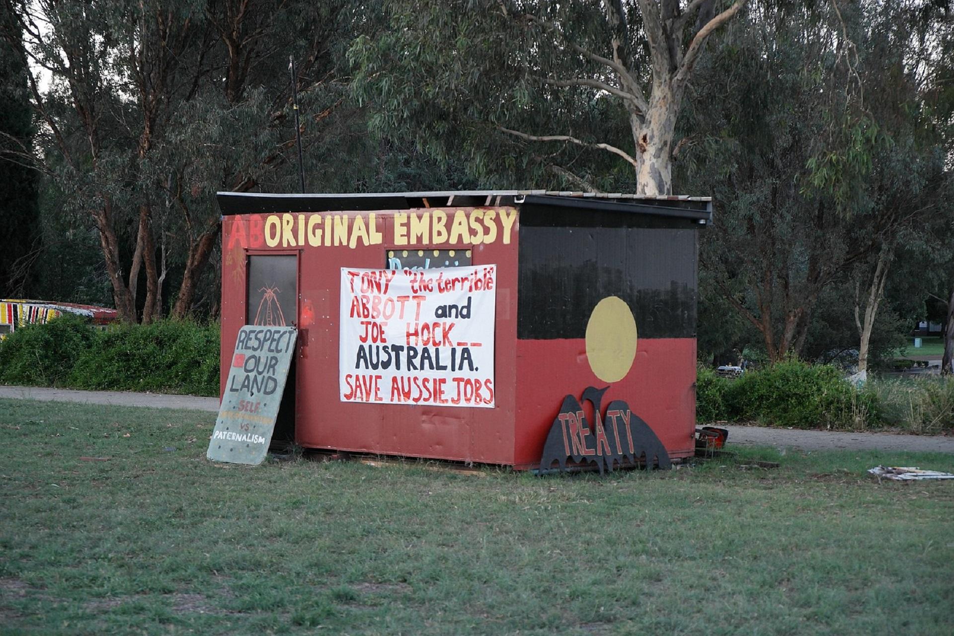 A long view of the rebuilt Aboriginal Embassy, painted in traditional red and black with a gold spot, bearing signs calling out various political themes.