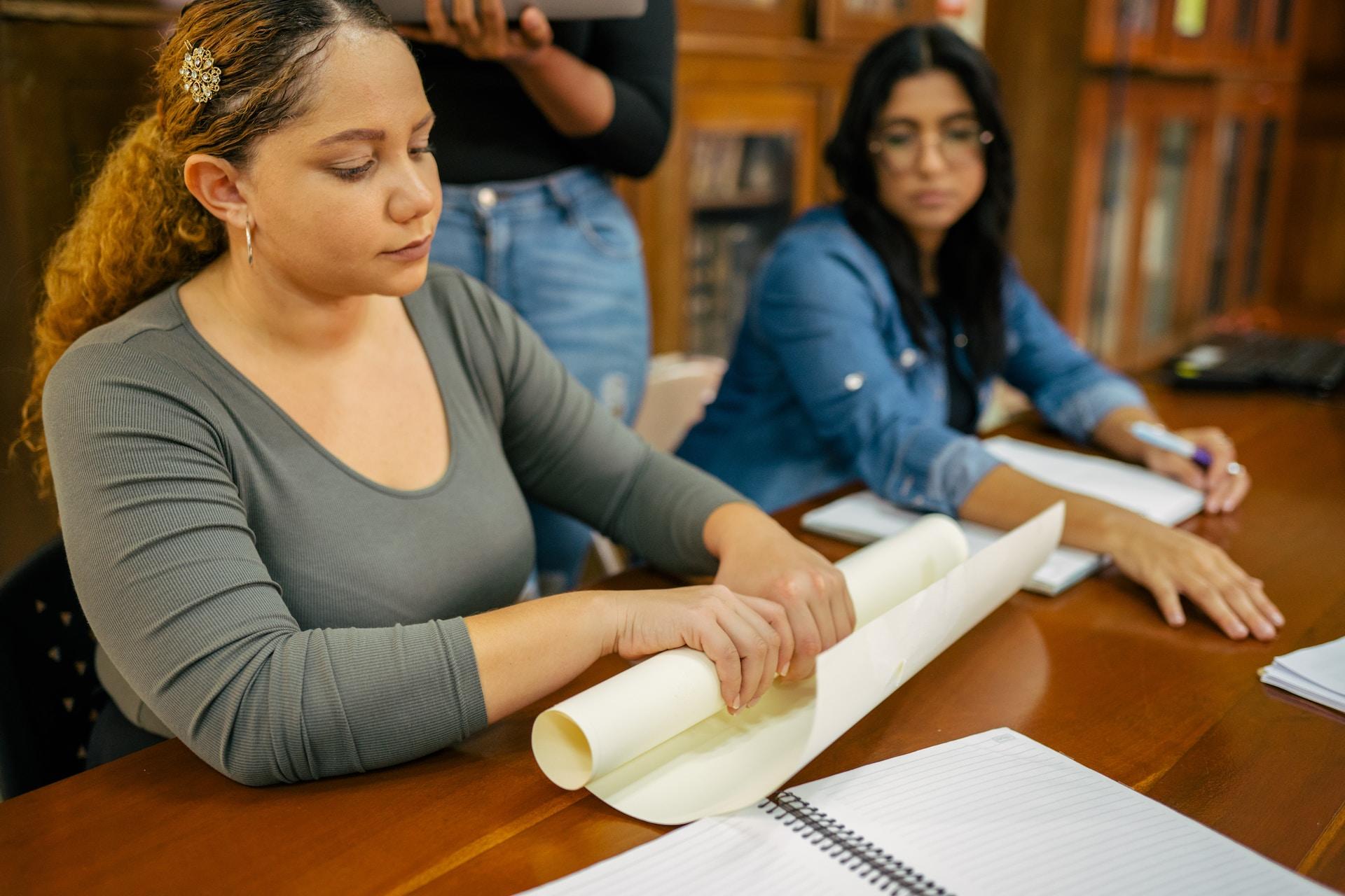 Two people sit next to each other at a long wooden table. The person in the green shirt rolls up a sheet of poster board while the person next to them, in blue, looks on.