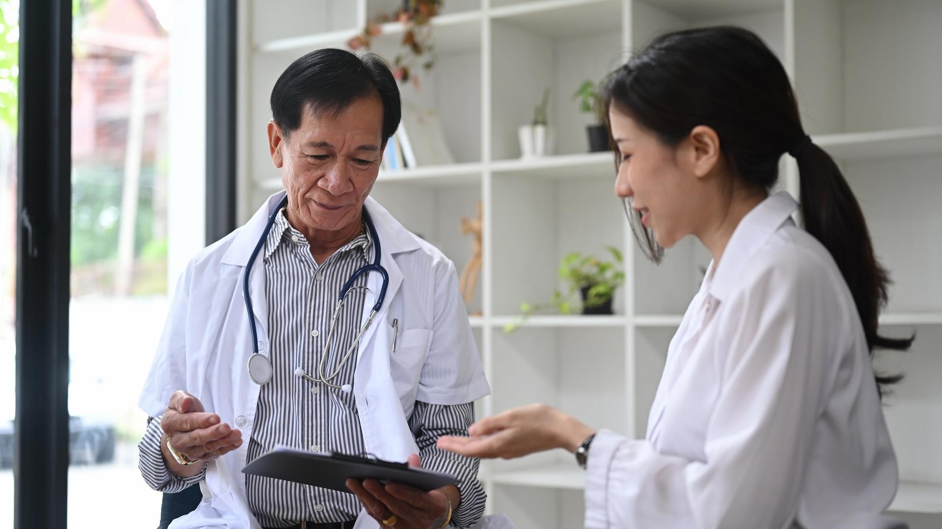 Two people dressed as doctors, one with a stethoscope around their neck and holding a patient chart, stand in front of a white shelving unit that features a small green plant.