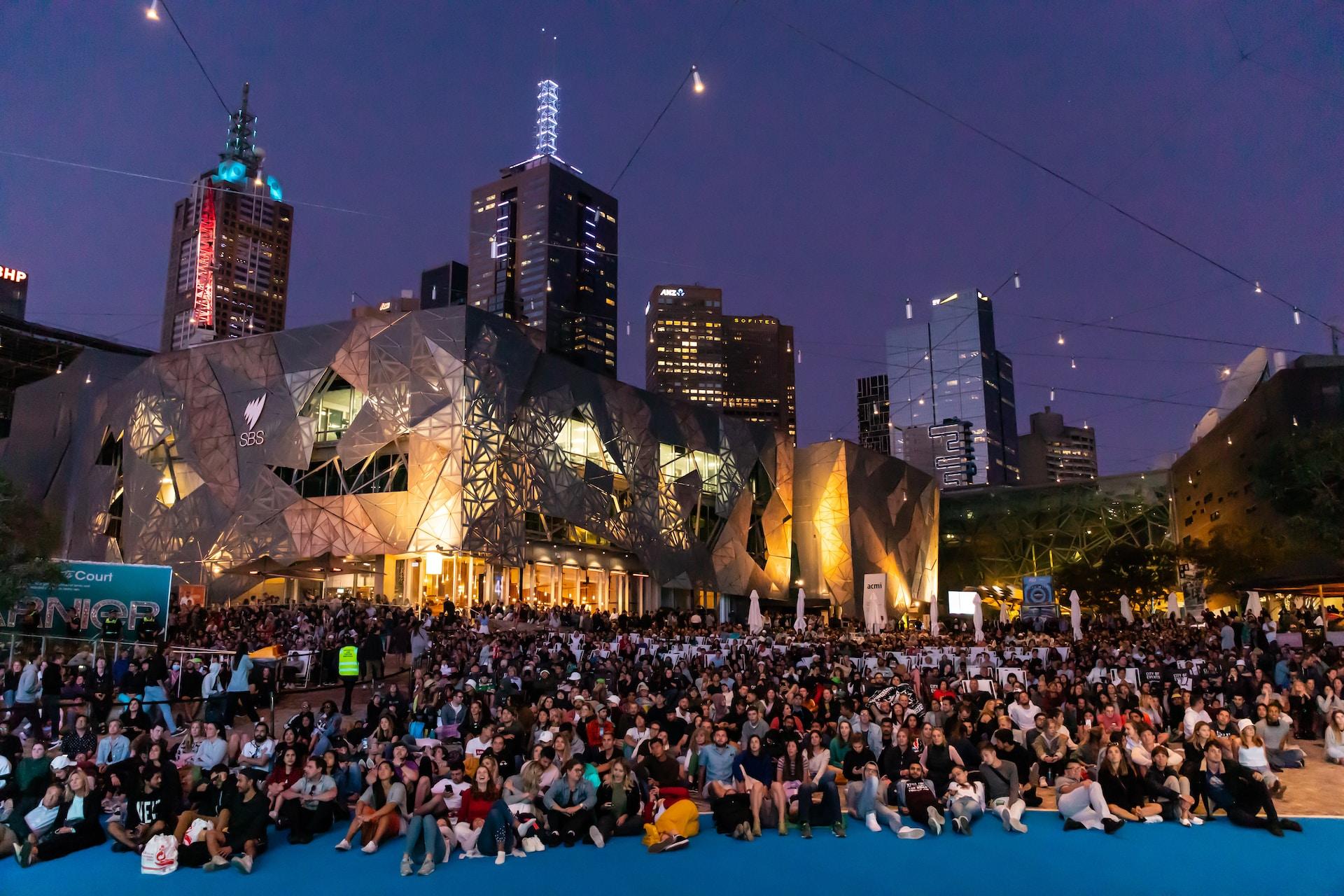 A large crowd sits on a Melbourne beach at night, with the city lit up in the background and the sky appearing purple.
