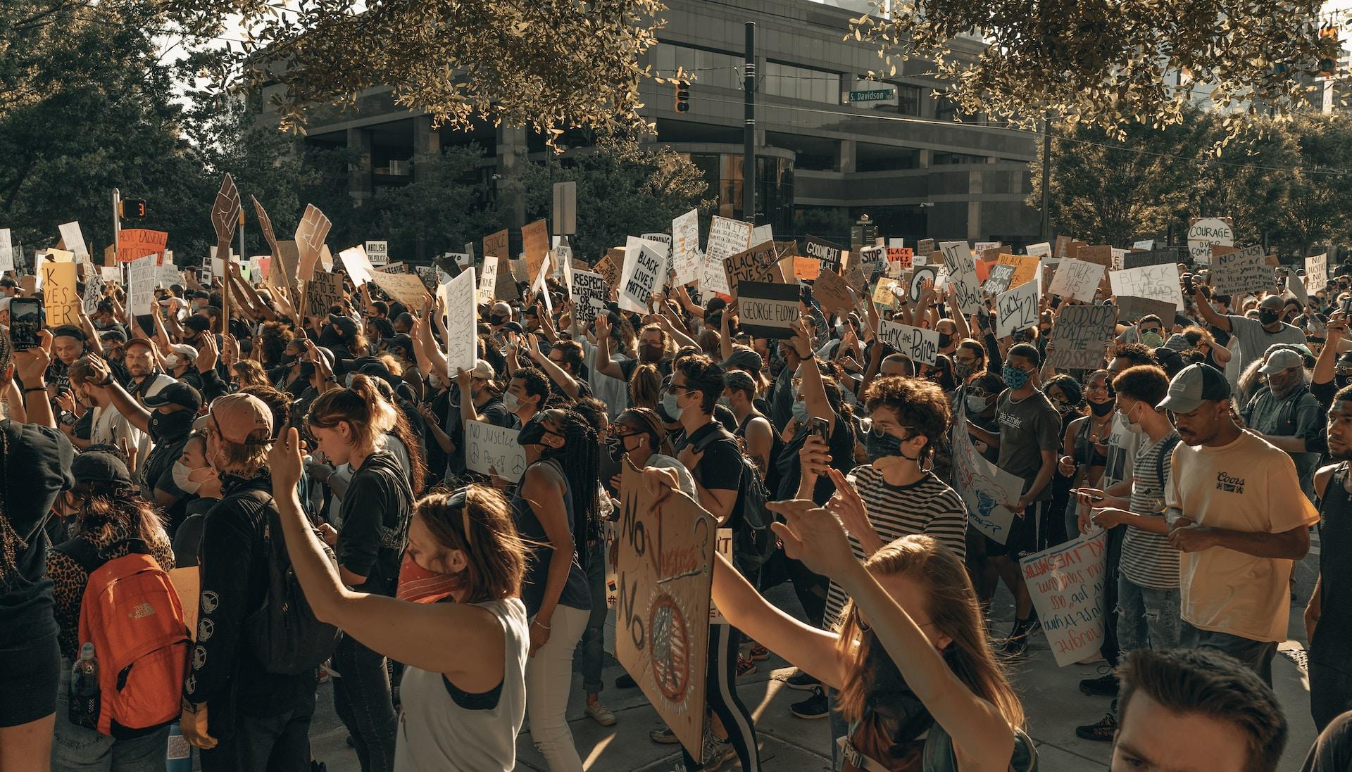 A large crowd of people, with some wearing face masks and holding protest signs walk down a street in a muted light that makes the photo seem sepia-toned.