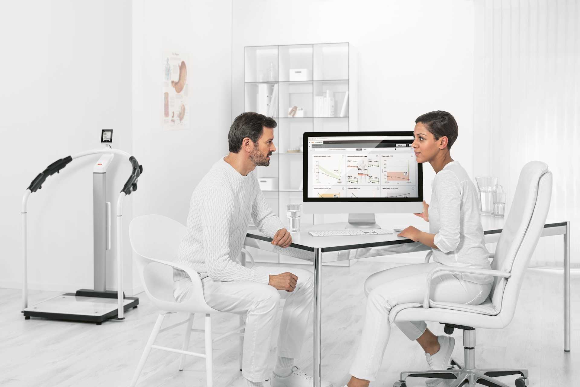 A patient wearing white sits in a very white diagnostic room talking with a technician who is also wearing white.