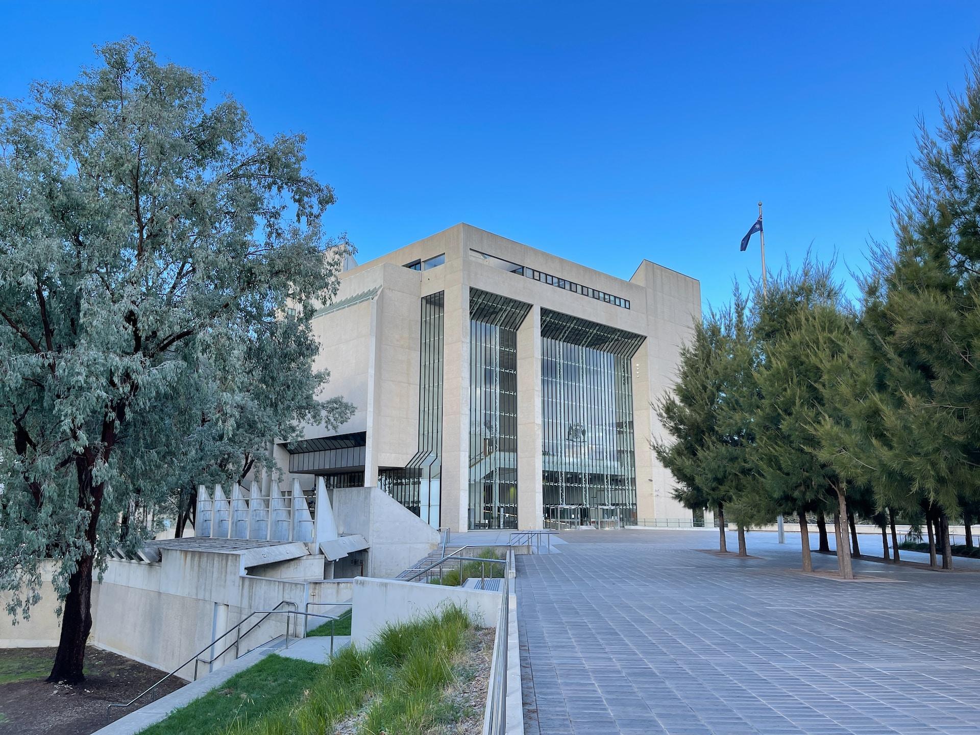 The Australian High Court building showing its high glass facade built into white stone construction, with a long driveway leading to it, under a clear blue sky.