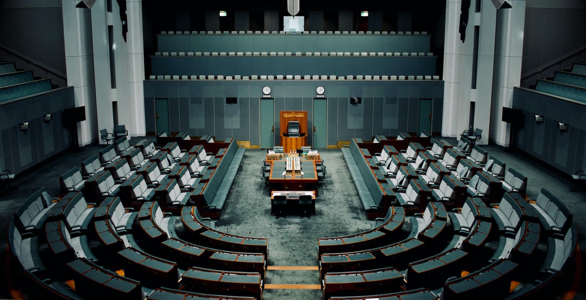 The House chamber of the Australian Parliament done in green tones with white seat cushions arranged in a horseshoe around a wooden table.