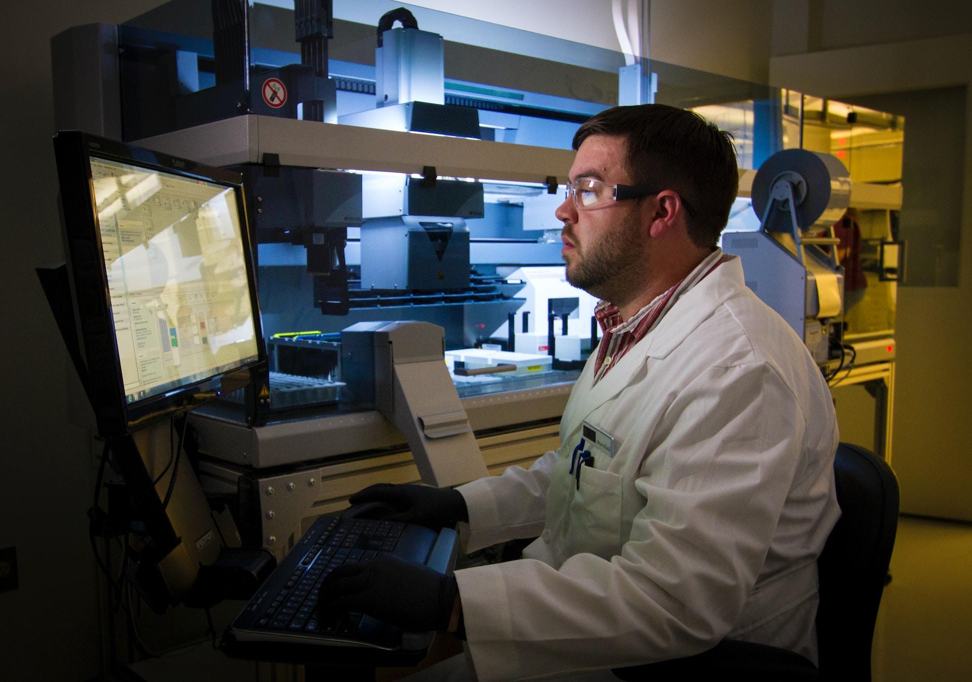 A person sits in front of a computer monitor in a darkened room as they assess a patient's diagnostic screening while wearing a white lab coat and safety goggles. 