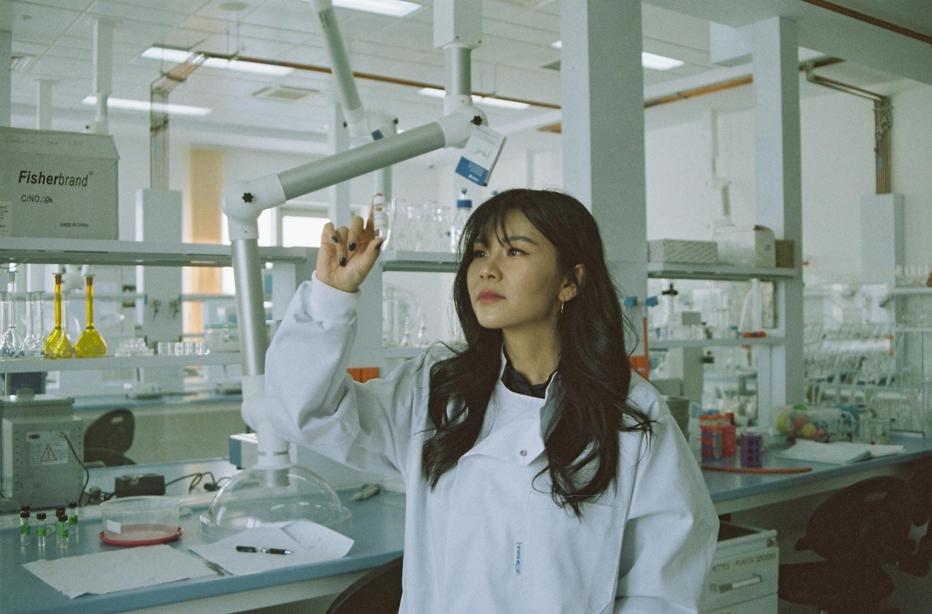 A person wearing a white lab coat stands in a laboratory, holding up a clear glass vial and inspecting its contents. 