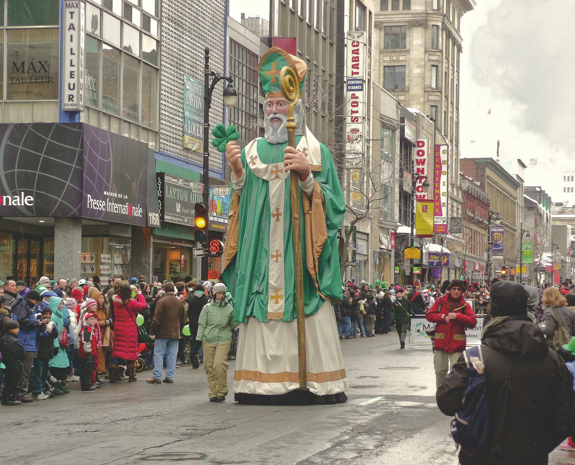 A parade in Ireland with crowds of people lining the street as a large religious statue bearing a crown and dressed in a green hassock goes by.