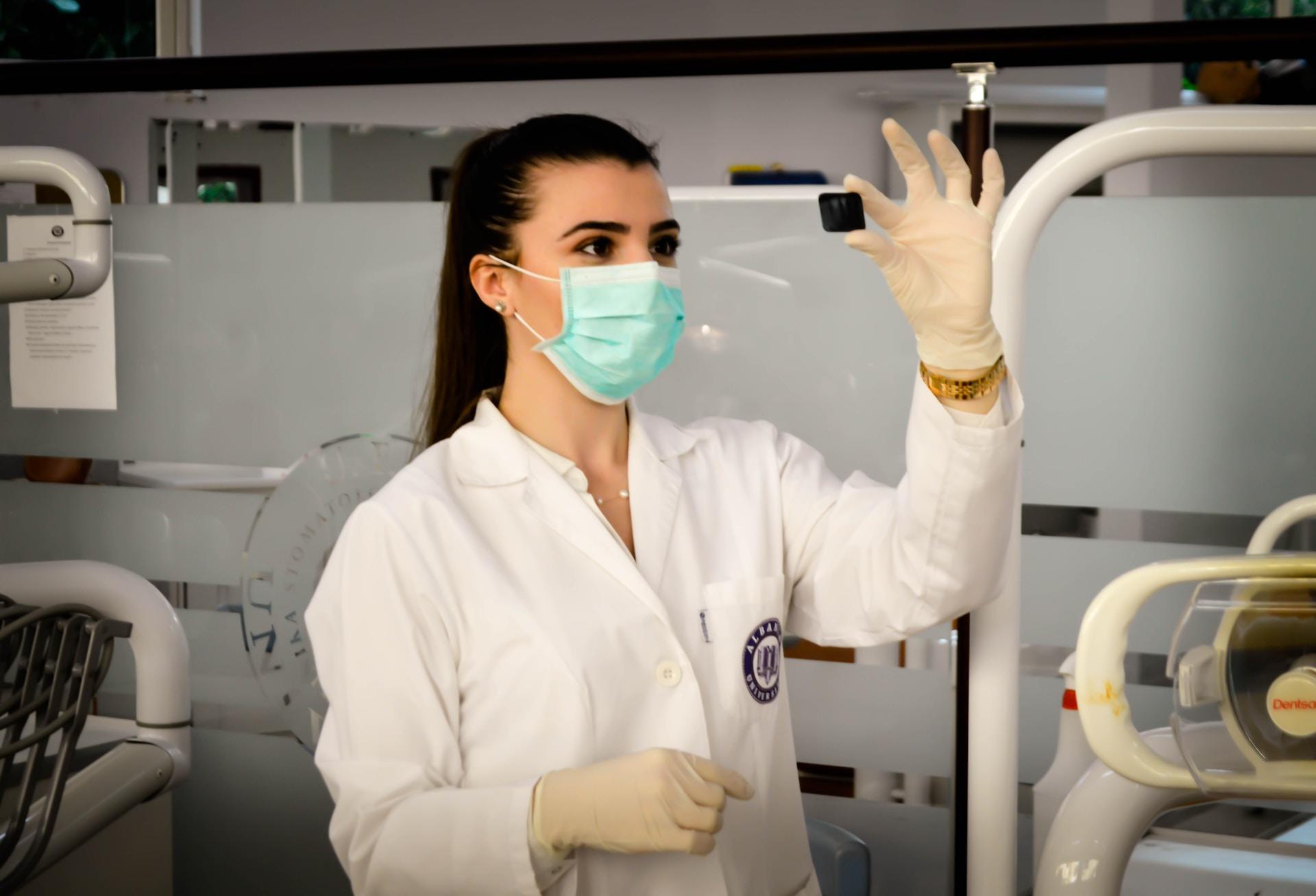 A person wearing a white lab coat and a blue face mask stands in a laboratory, holding up a medical slide and peering intently at it.