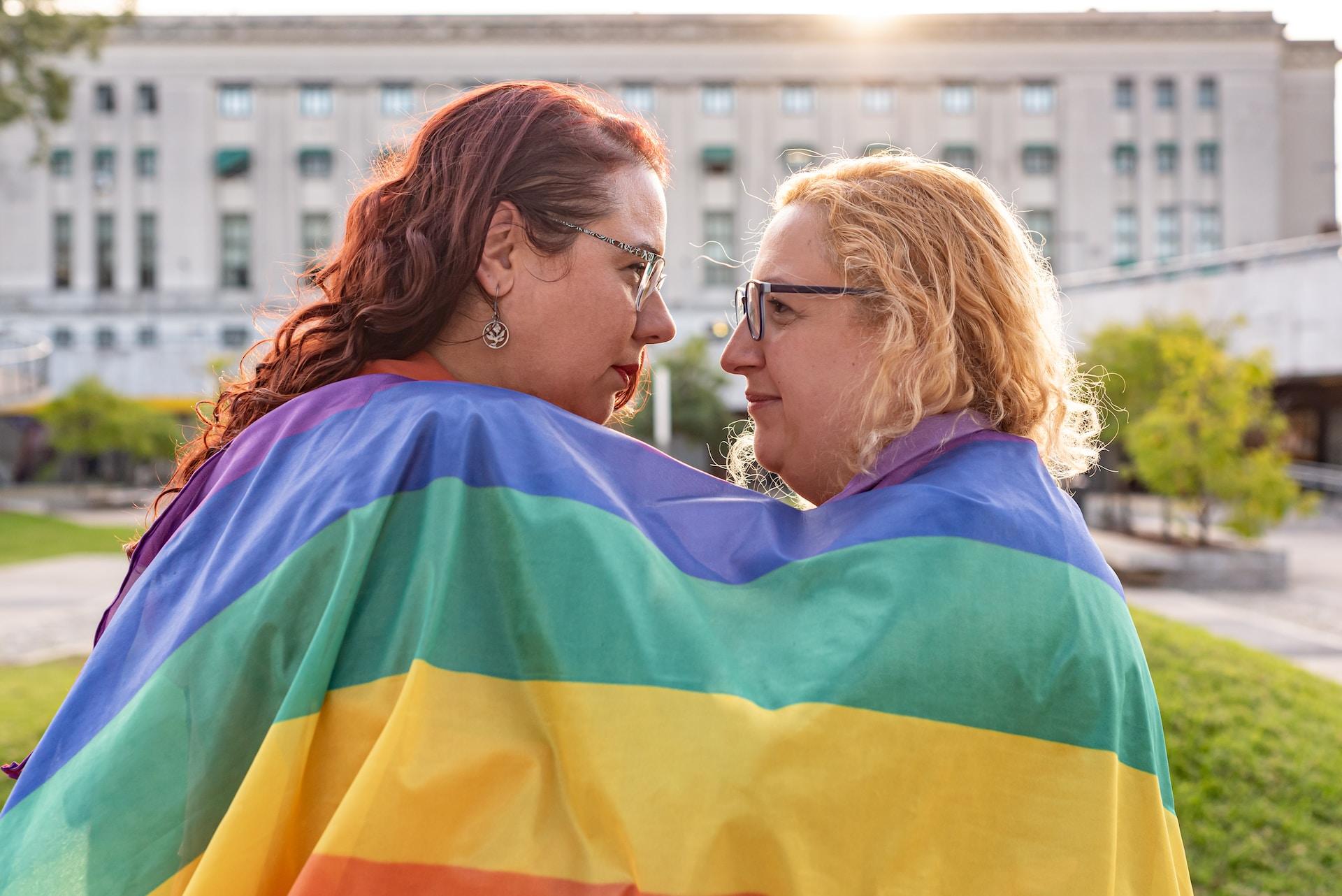 Two people draped in a rainbow flag stand face to face against the backdrop of an official building on a sunny day.