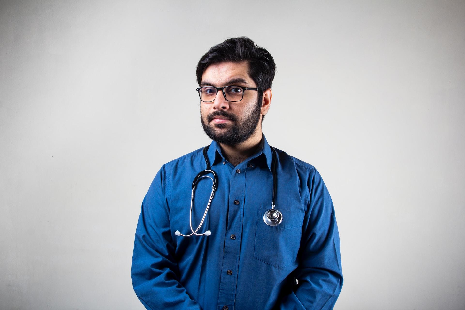A person wearing a blue shirt and dark-framed glasses bears a stethoscope around their neck stands against a plain white background and looks at the camera questioningly.