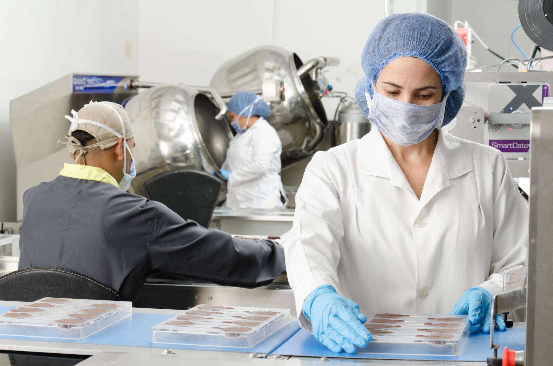 Three lab technicians, two wearing white lab coats and one wearing a slate grey coloured on, work on various tasks in a laboratory. They are all wearing safety gear such as nitrile gloves, splash goggles, face masks and hair nets.