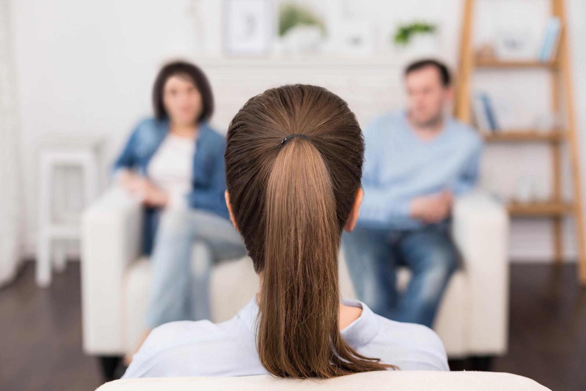 A person in a light grey top and their brown hair tied back faces two adults sitting on the white sofa opposite them in a brightly lit room.