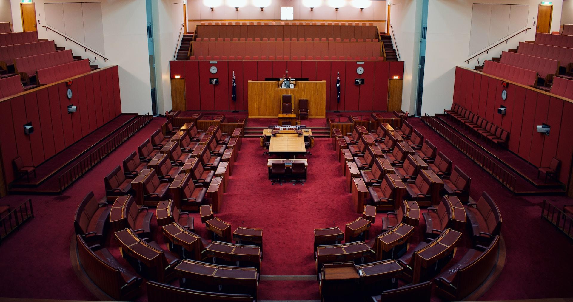 A long view of the Australian Parliament Senate chamber with its red carpet and wooden benches arranged in a semi-circle and spectator galleries lining the walls.